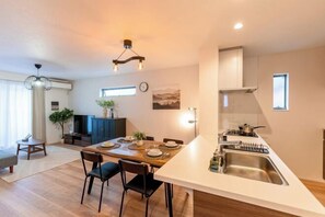 Eine moderne Küche mit Kochinsel, ein Essbereich mit Holztisch und -stühlen sowie ein Wohnbereich mit Sofa und Pflanze.