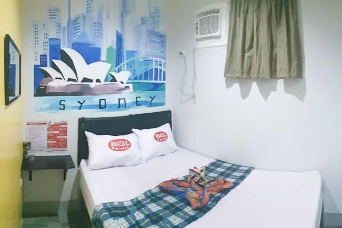 A hotel room with a bed, a wall with a cityscape and 'SYDNEY' text, a window with curtains, and a nightstand with a lamp.