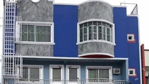 A two-story building with a blue and gray facade, a balcony with a metal railing, and a circular window on the upper floor.