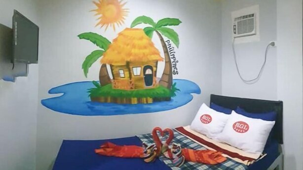 A room with a bed, a wall mural of a tropical scene, and a television mounted on the wall.