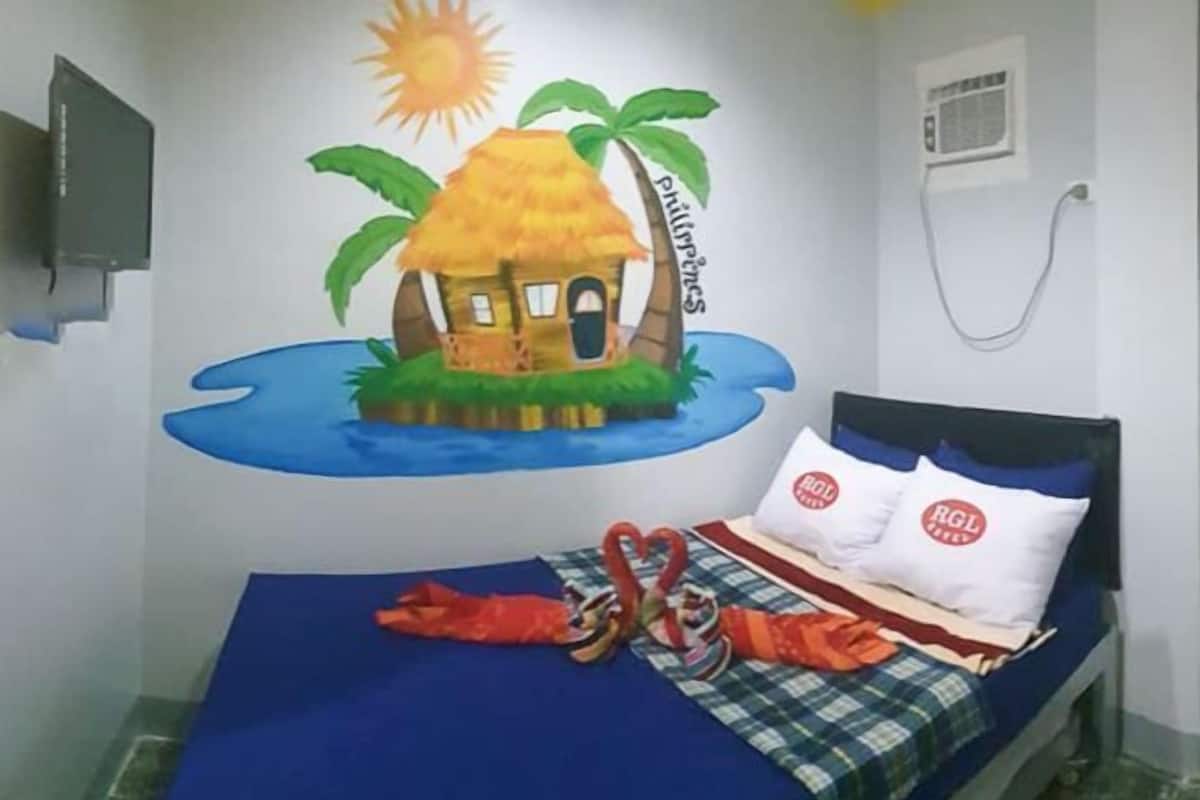 A room with a bed, a wall mural of a tropical scene, and a television mounted on the wall.