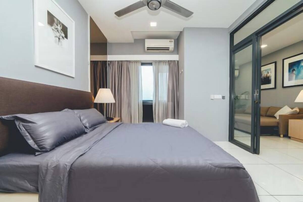 A modern hotel room with a bed, a ceiling fan, an air conditioning unit, a nightstand, a lamp, a window with curtains, and a sliding glass door leading to a balcony.