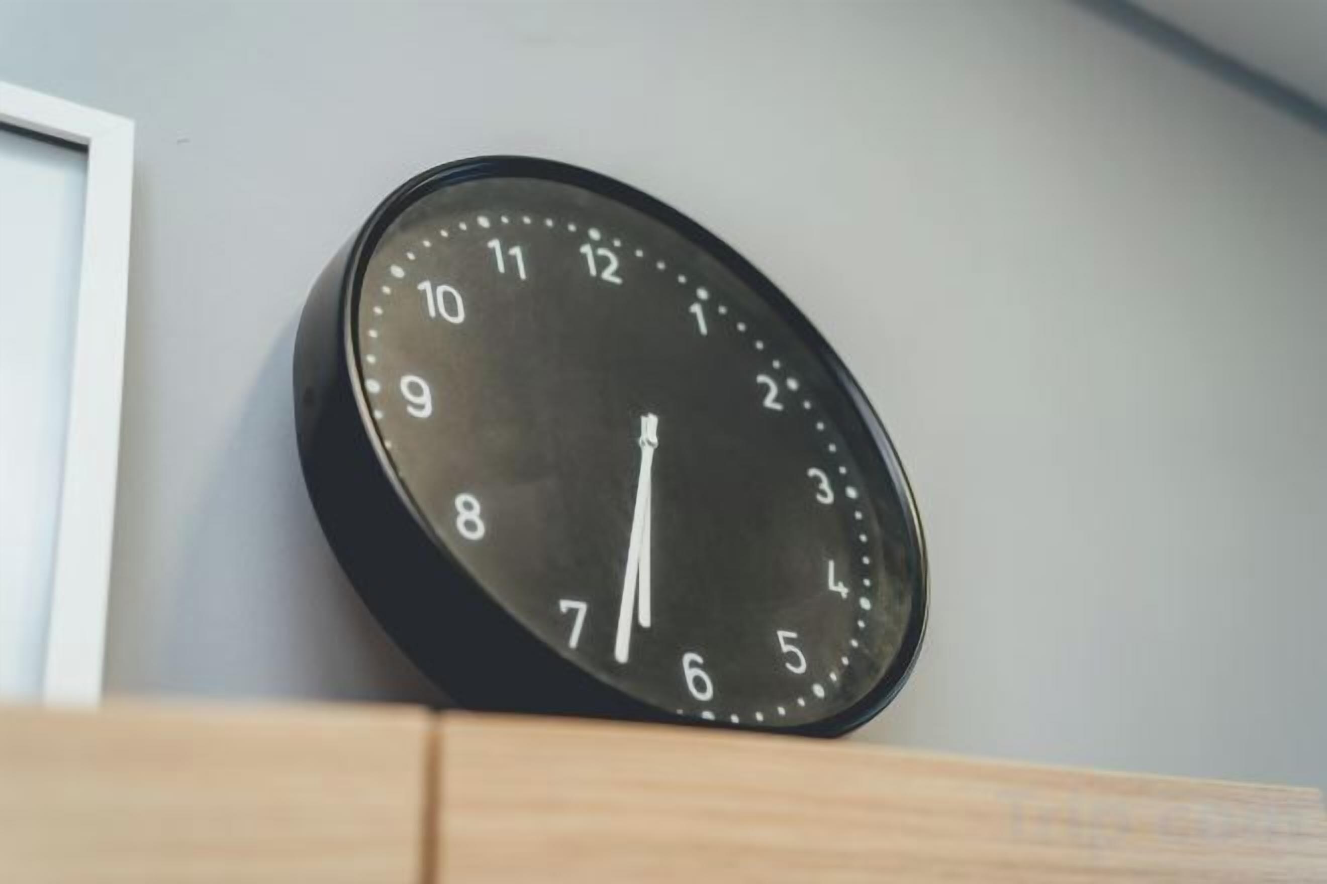 Eine runde Wanduhr mit weißen Zahlen und Zeigern, die an einer Holzfläche vor einer schlichten Wand angebracht ist.