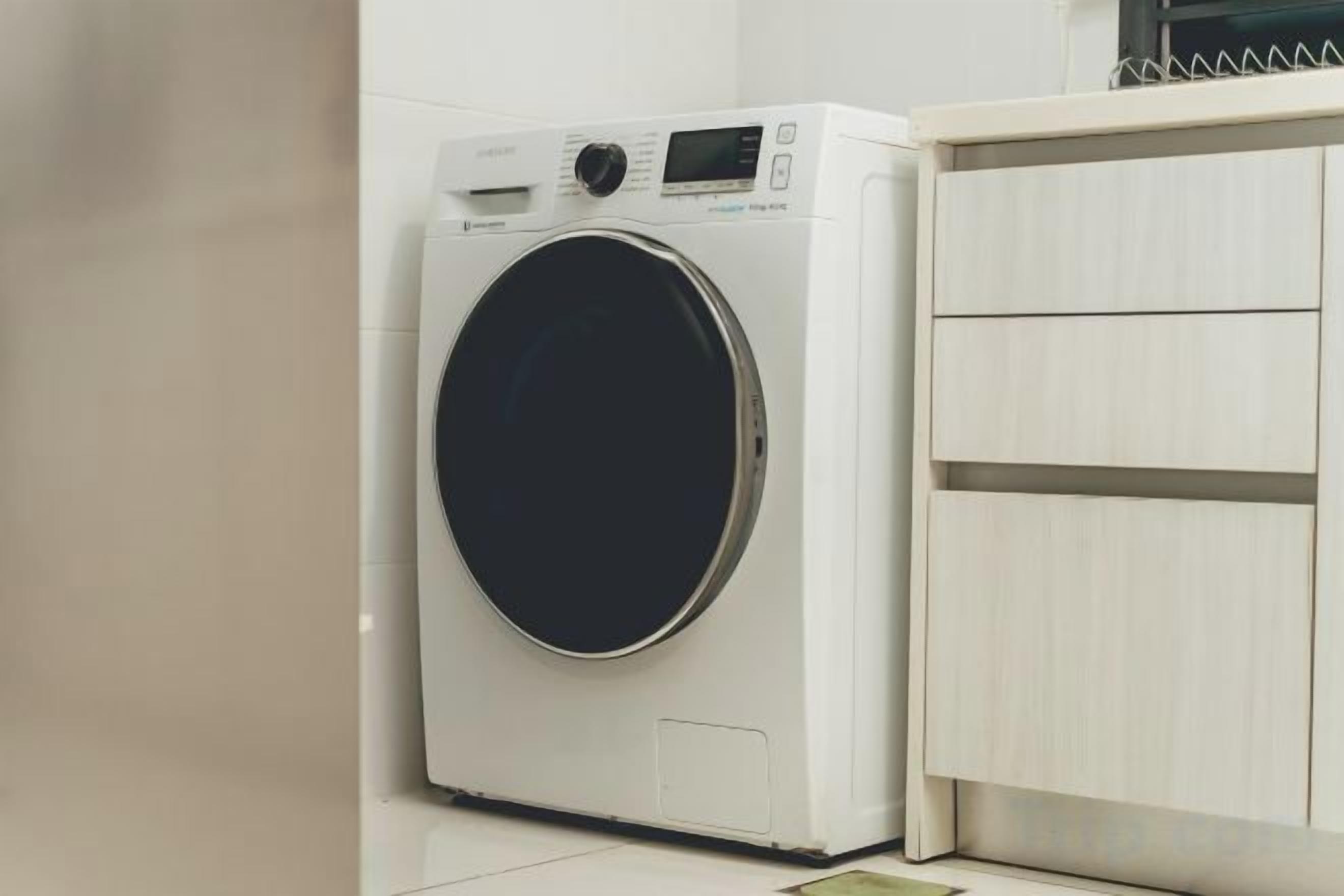 A front-loading washing machine in a kitchen with a wooden cabinet.
