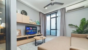 Una habitación de hotel moderna con cama, televisor sobre un mueble de madera, una mesita redonda pequeña, ventilador de techo y un ventanal grande con cortinas.