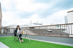 A person sitting on a folding chair with a laptop on a rooftop terrace with a view of buildings and a railing.