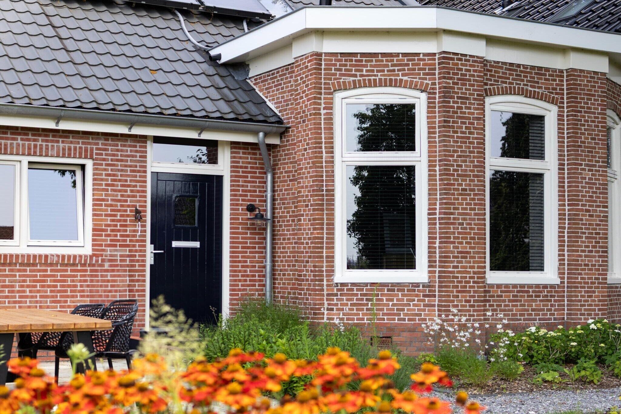 Une maison d’angle en briques avec une porte noire, deux fenêtres et une table en bois avec des chaises.