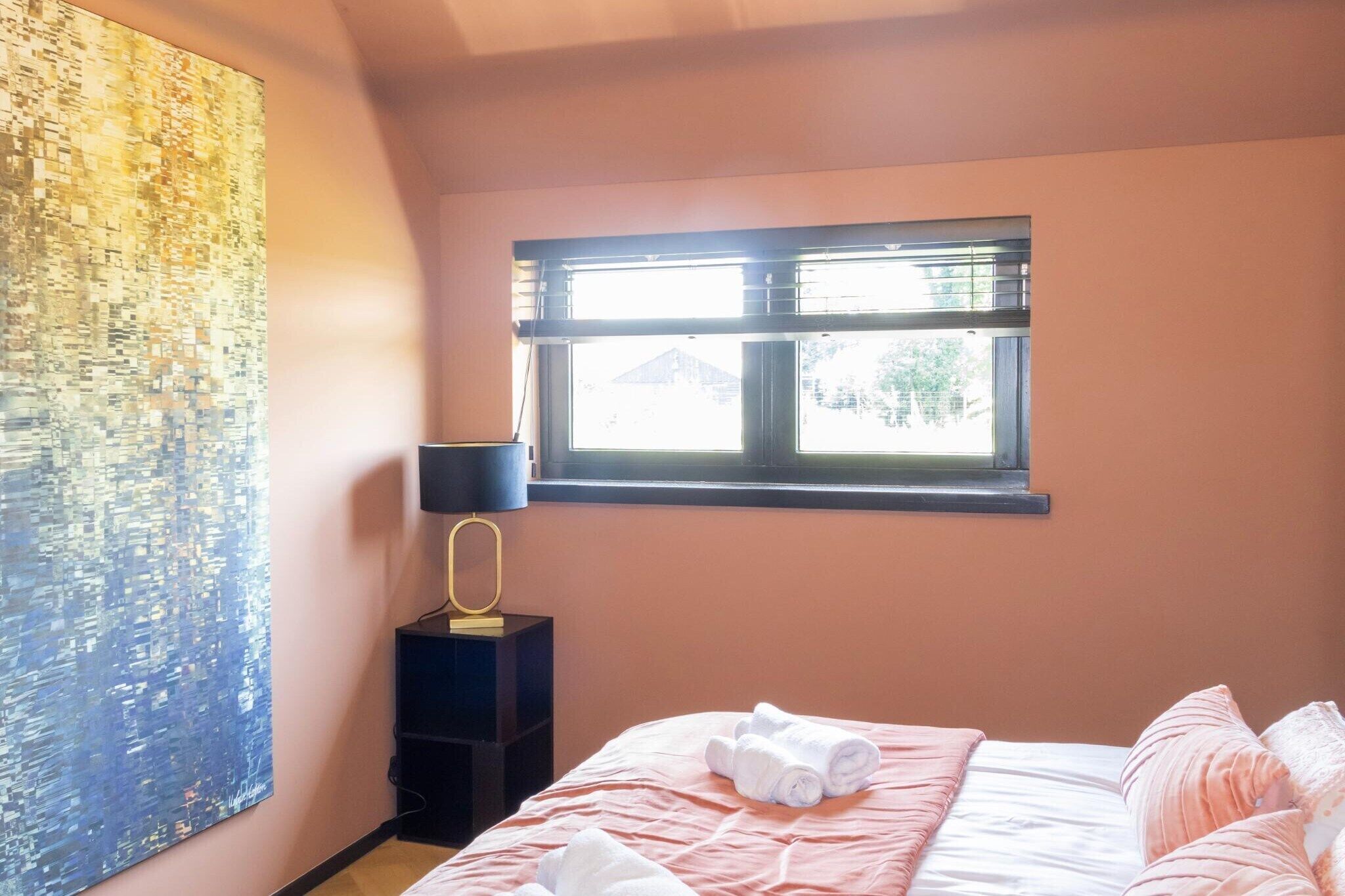Une chambre à coucher avec un lit, une fenêtre, un tableau décoratif au mur et une table de chevet avec une lampe.
