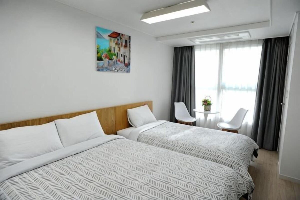 A hotel room with two beds, a painting on the wall, a small table with a plant, and a view of the outdoors through large windows.