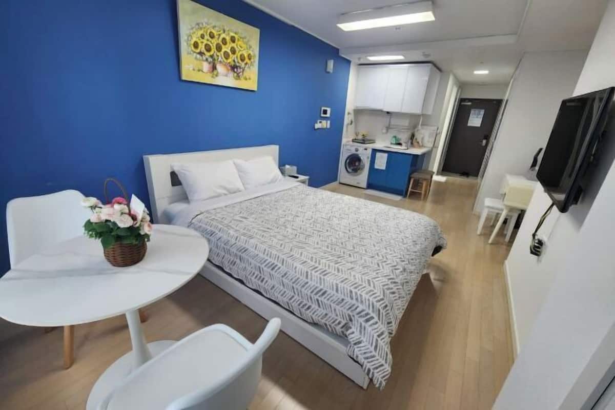 A bedroom with a bed, a small table with a basket, a chair, a kitchenette, and a TV.