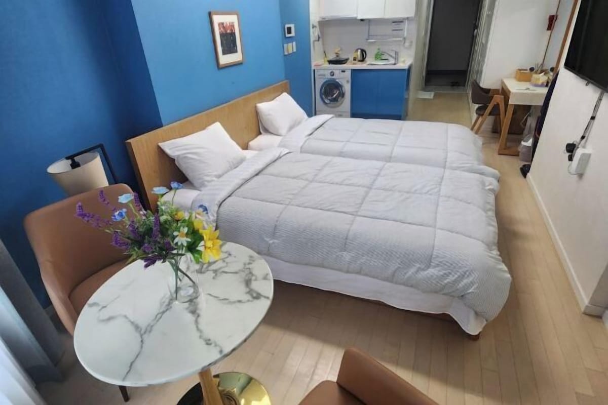 A hotel room with a bed, a small table with a vase of flowers, a chair, and a washing machine in the background.