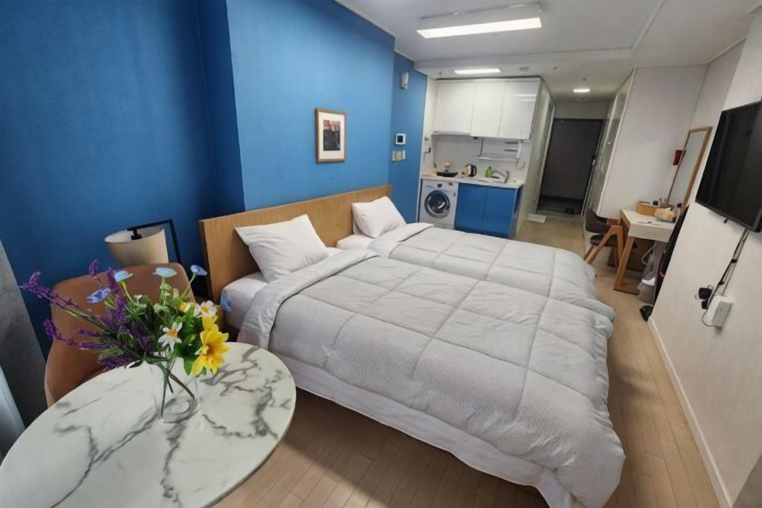 A hotel room with a bed, a round table with a vase, a desk with a chair, and a kitchenette in the background.
