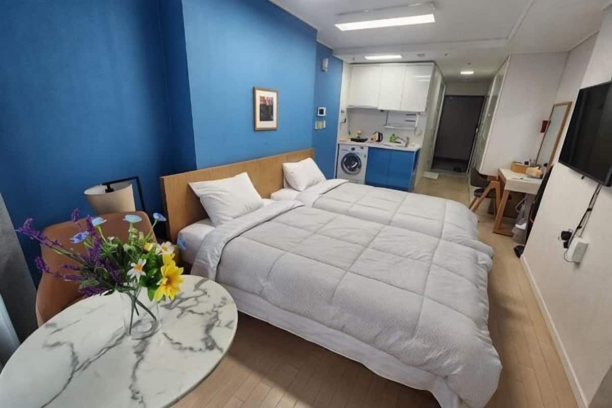 A hotel room with a bed, a round table with a vase, a desk with a chair, and a kitchenette in the background.