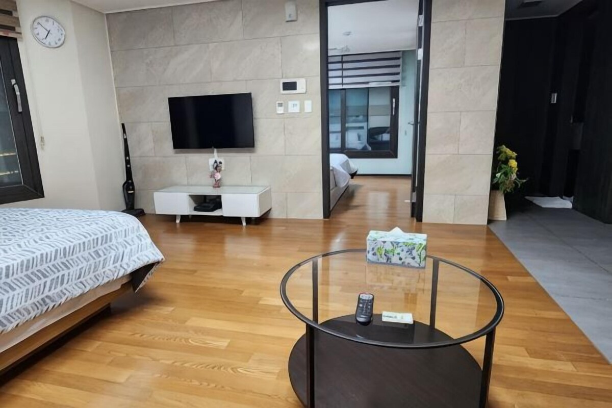 A modern hotel room with a bed, a TV, a small white cabinet, a glass coffee table, and a view of the hallway.