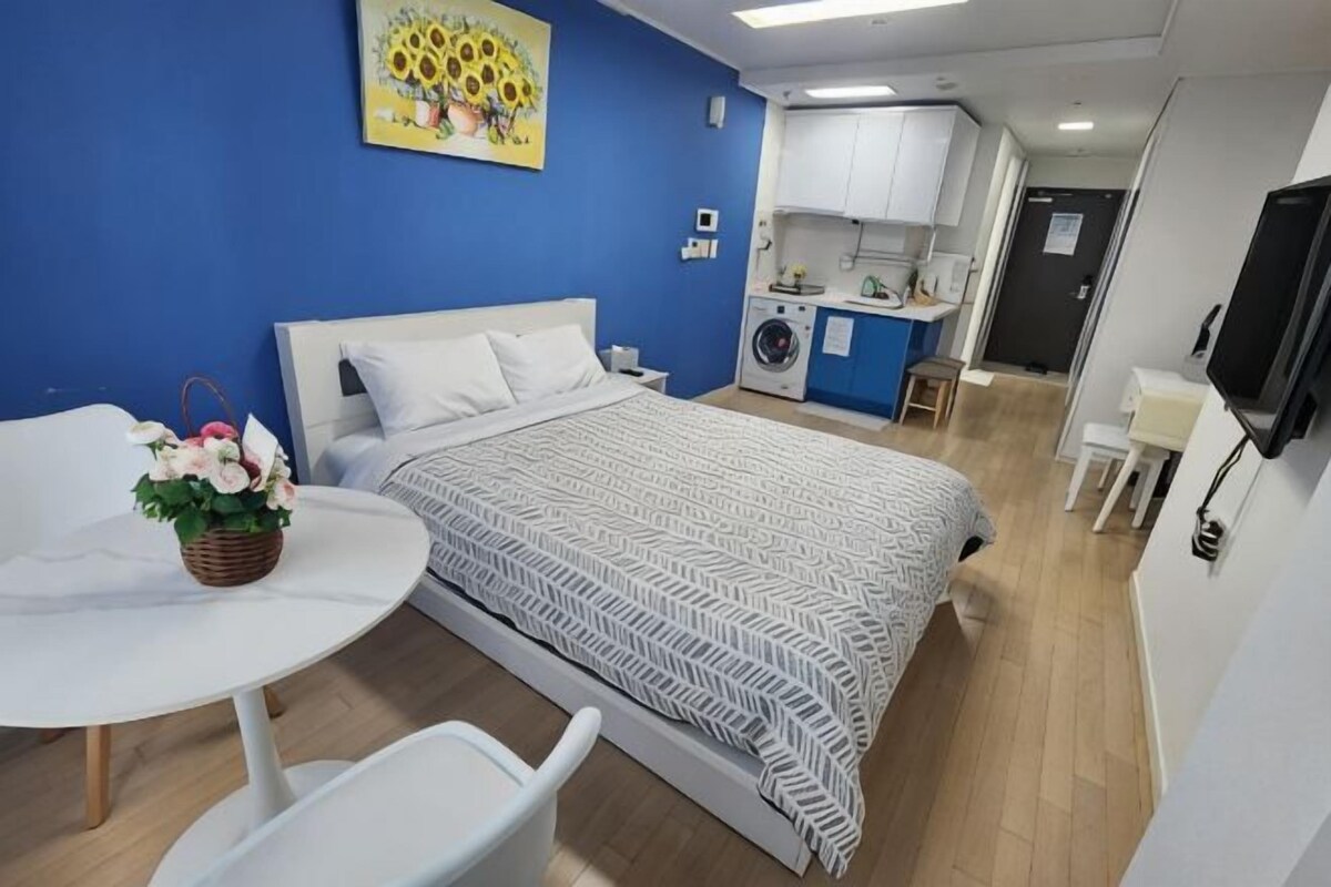 A bedroom with a bed, a small table with a basket, a white chair, and a kitchenette in the background.