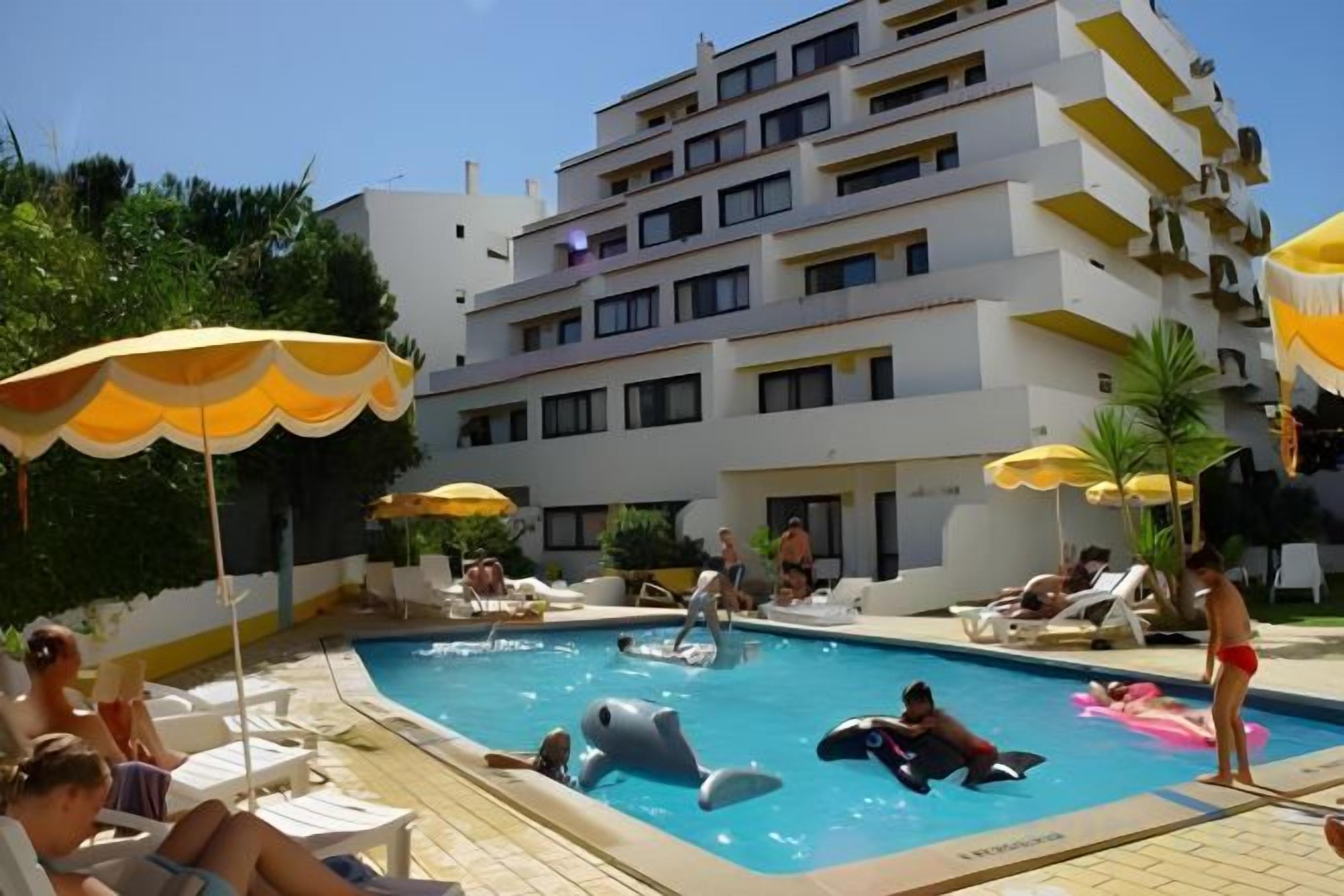 Piscina com espreguiçadeiras e guarda-sóis, cercada por um edifício de vários andares.