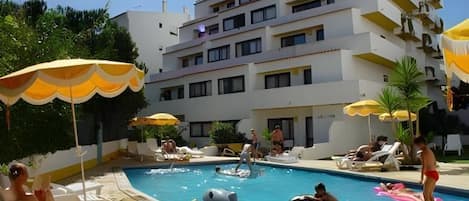 Ein Schwimmbecken mit Liegestühlen und Sonnenschirmen, umgeben von einem mehrstöckigen Gebäude.