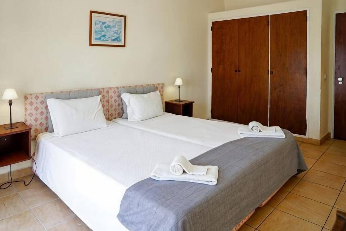 Ein Doppelbett mit zwei Kissen, ein Nachttisch mit Lampe, ein Holzschrank und ein gerahmtes Bild an der Wand.