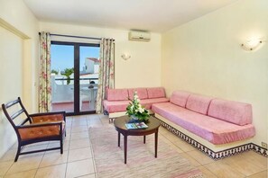 Un salon avec un canapé rose, une chaise en bois, une table basse et une vue à travers une porte vitrée.
