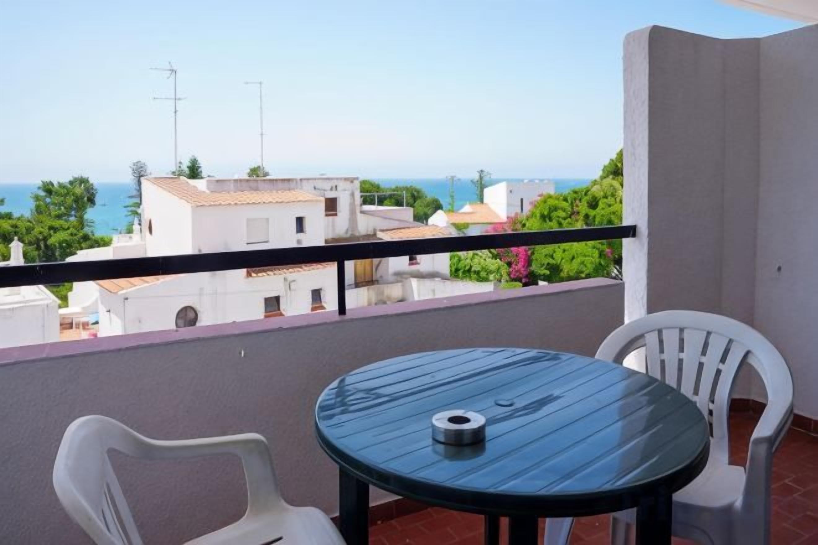 Un balcon avec une table ronde et deux chaises, offrant une vue sur la côte, avec des bâtiments et de la végétation.