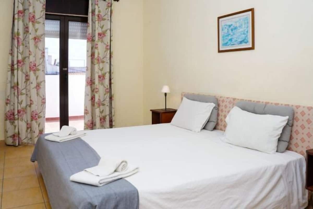 Ein Hotelzimmer mit Bett, Nachttisch, Lampe, einem gerahmten Bild an der Wand und Blick durch ein mit Vorhängen drapiertes Fenster.