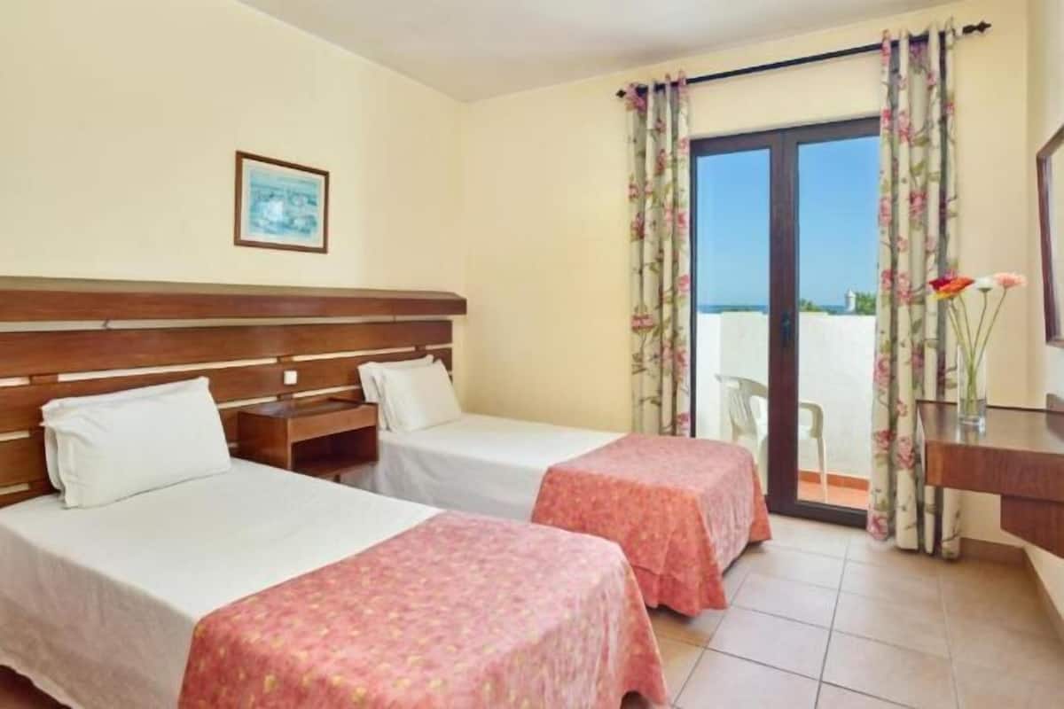 Ein Hotelzimmer mit zwei Betten, einem Holzkopfstück, einem Schreibtisch und Blick durch eine Glastür.