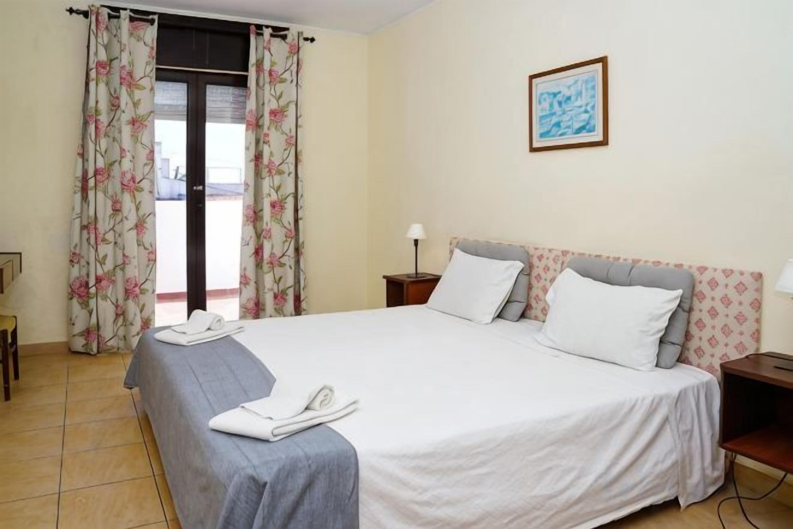 Quarto de hotel com cama, cortinas, mesa de cabeceira, abajur e um quadro na parede.