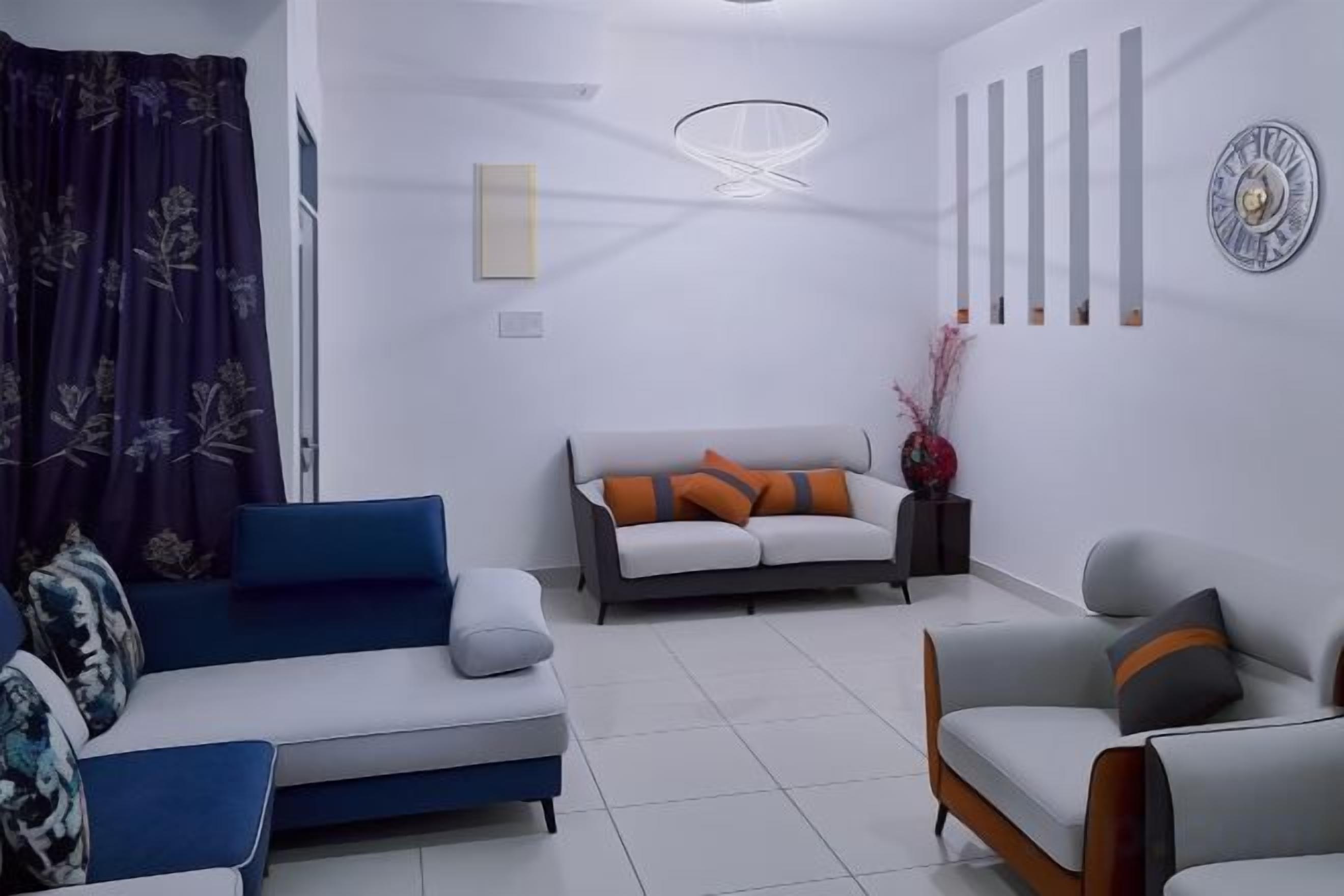 A modern living room with a blue sofa, a grey sofa with orange cushions, and a grey armchair. There is a white coffee table, a decorative vase with flowers, and a wall-mounted light fixture.