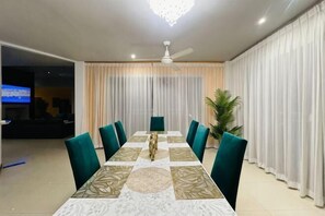 A dining area with a long table, chairs, curtains, a ceiling fan, and a TV in the background. - The Ace 6 Bedrooms 6 Bathrooms Villa With Free Bills (Pattaya)
