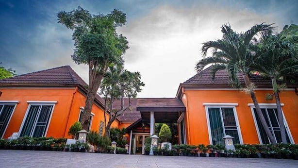 A two-story building with a red-tiled roof and orange walls, surrounded by palm trees and potted plants. - The Ace 6 Bedrooms 6 Bathrooms Villa With Free Bills (Pattaya)
