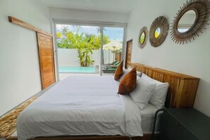 A bedroom with a bed, a desk, a chair, a large window, and a view of a pool and greenery. - Villa Kurma, 2 bedrooms (Koh Samui)