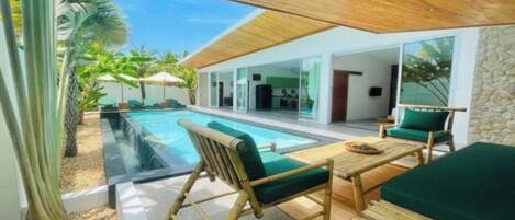 A modern outdoor pool area with a wooden deck, green cushioned seating, and a stone wall backdrop.