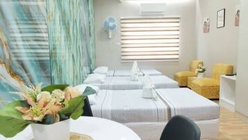 A hotel room with two beds, a small table with a vase, a fan, and a window with blinds.