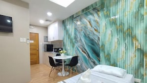 Quarto de hotel com cama, escrivaninha, cadeira, mesa pequena com um vaso e área de cozinha ao fundo.