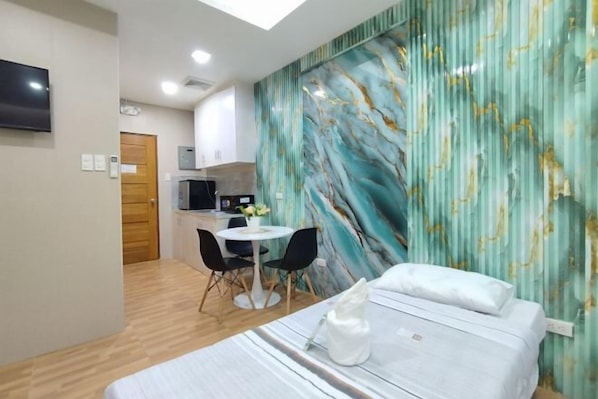 A hotel room with a bed, a desk, a chair, a small table with a vase, and a kitchen area in the background. - Townhouse Oak Casa Barcelona (Iloilo City)