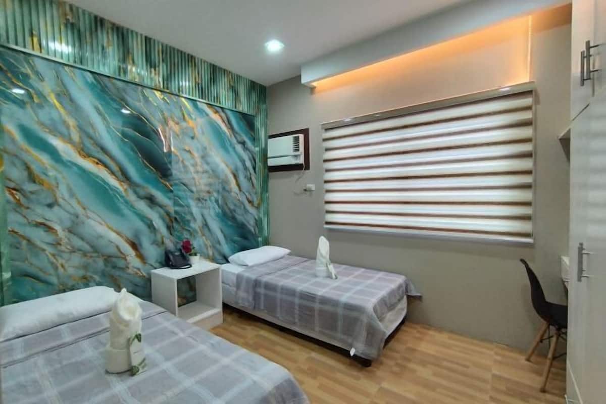 Habitación de hotel con dos camas, una pared de mármol, una ventana con persianas y una mesita de noche pequeña.