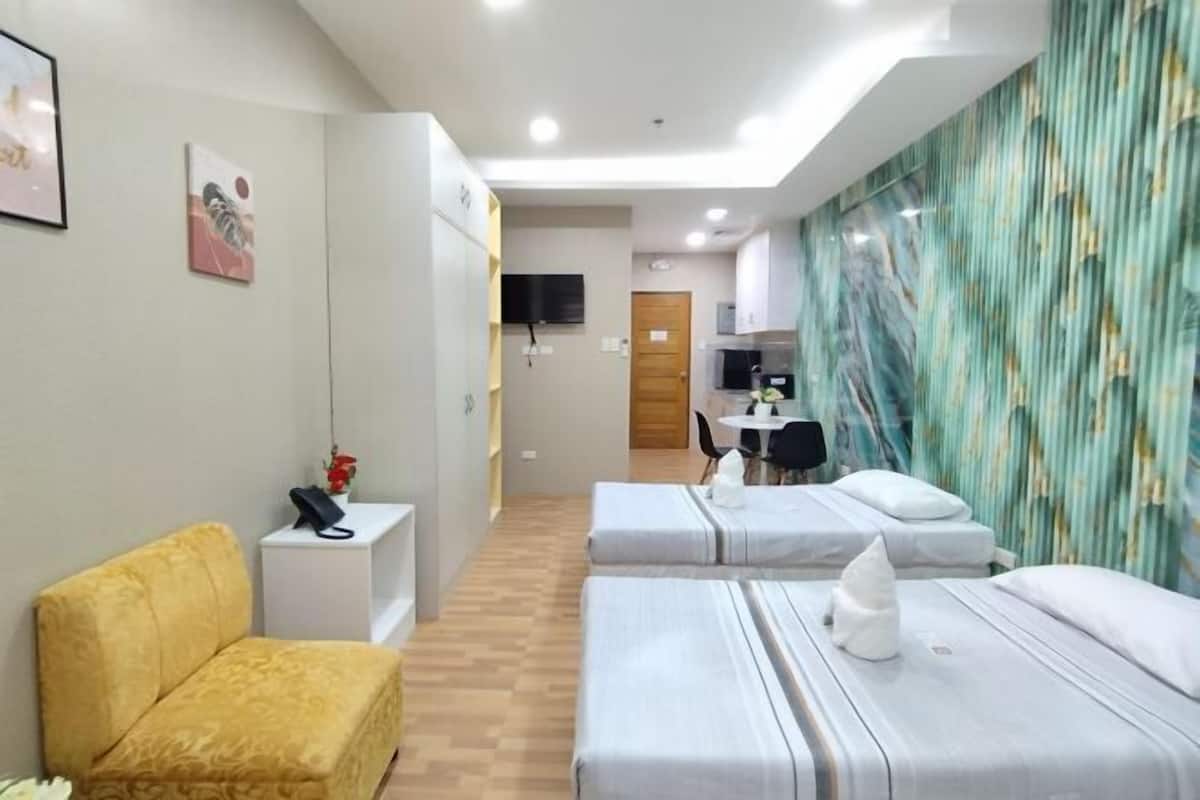 Habitación de hotel con dos camas, un sofá, una pequeña mesa, un televisor y una zona de cocina al fondo.