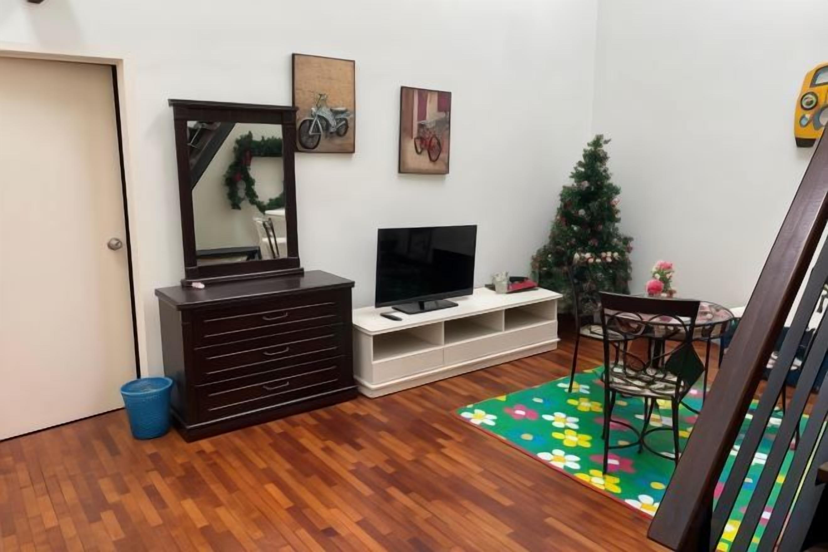 A room with a wooden floor, a dark-colored dresser with a mirror, a flat-screen TV on a white stand, a small Christmas tree, and a dining table with chairs.