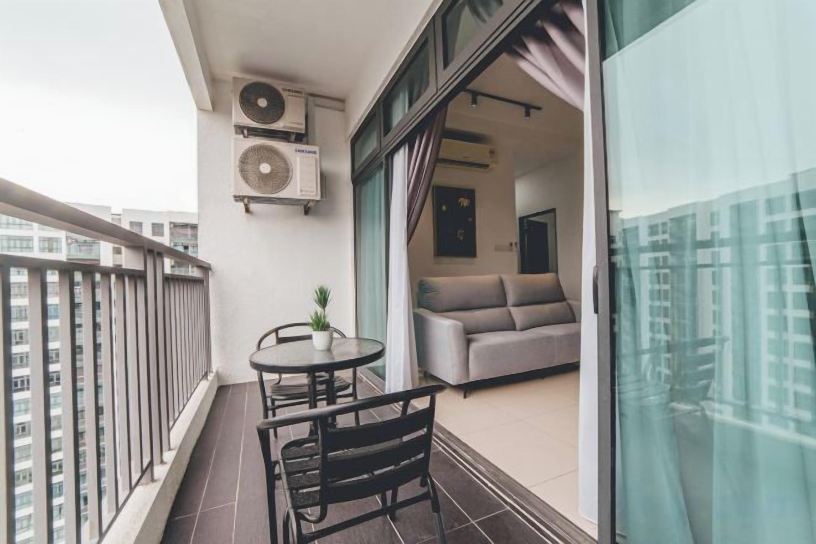 A balcony with a table and chairs, a sofa inside the adjacent room, and two air conditioning units mounted on the wall.