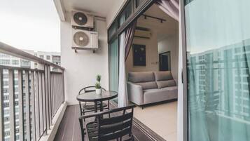 A balcony with a table and chairs, a sofa inside the adjacent room, and two air conditioning units mounted on the wall.