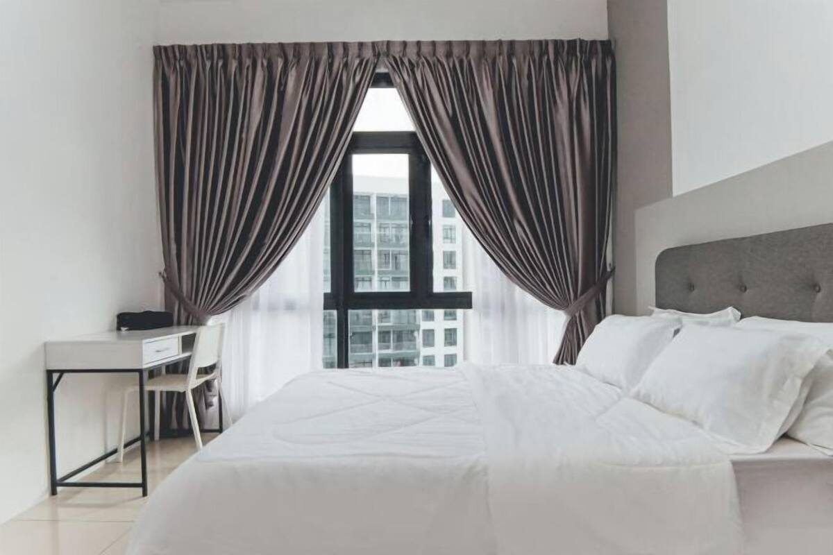 A modern hotel room with a large bed, a desk, and a chair, featuring a view of the city through large windows.