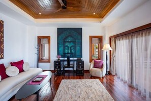 A living room with a sofa, a coffee table, a desk with a chair, a Buddha painting, and a large window with curtains. - Villa ZeniMo, Surin Beach, pool, 9 BR 18 guests (Phuket)