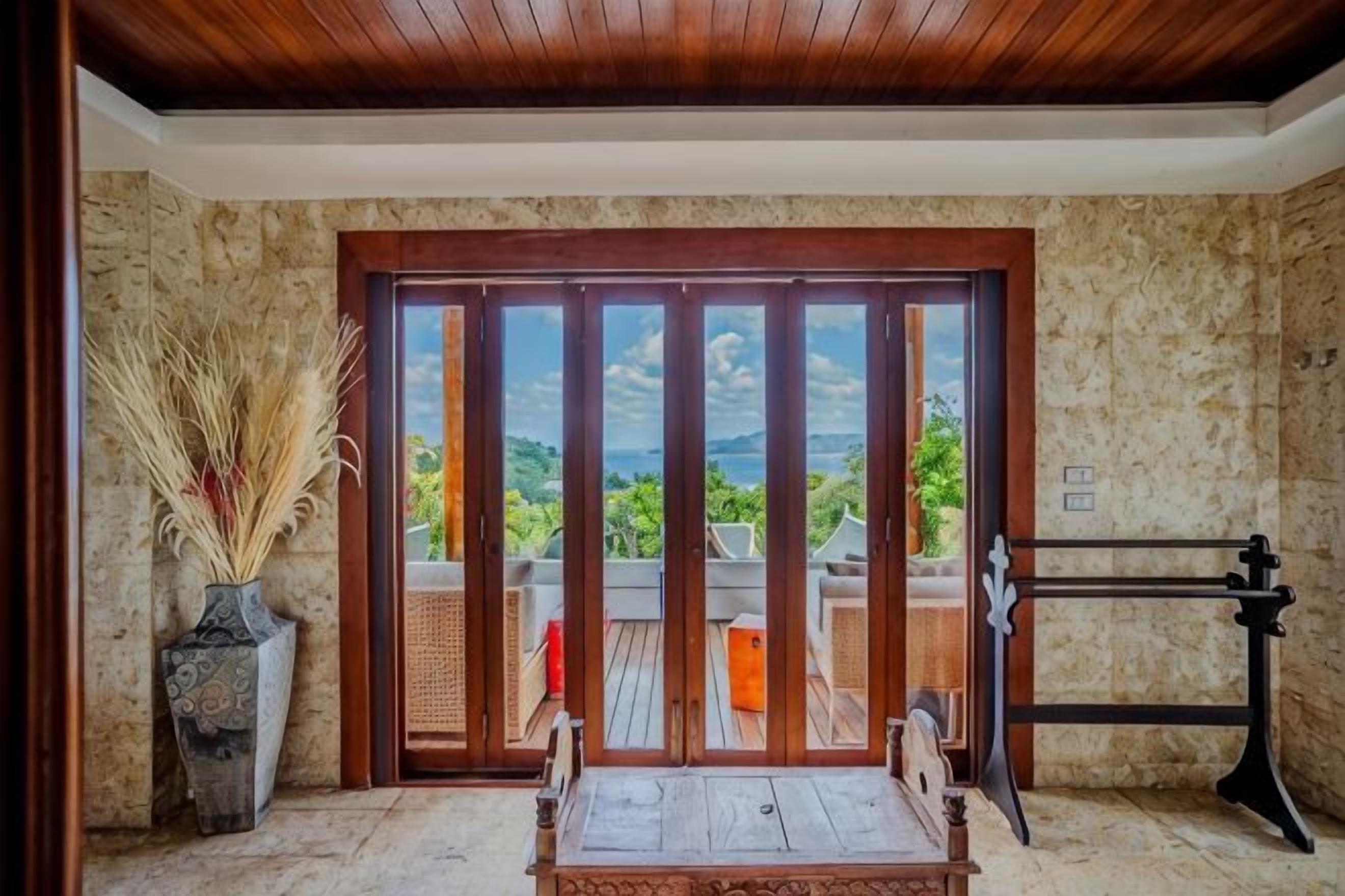 A room with a wooden ceiling, a large sliding door leading to a balcony, a table with chairs, and a decorative vase.