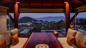 A room with a wooden table and two sofas, a view of a cityscape and mountains, and a ceiling with wooden beams. - Villa ZeniMo, Surin Beach, pool, 9 BR 18 guests (Phuket)