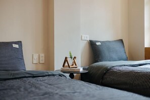Habitación de hotel con dos camas, cada una con ropa de cama a rayas, una mesita con un libro y una planta, y un enchufe en la pared.