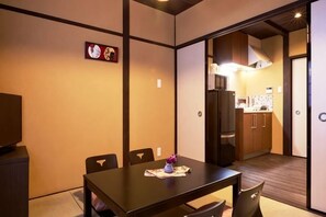 A room with a dining table, chairs, a TV, and a kitchen area visible through an open door.