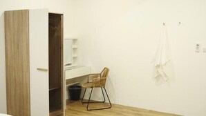 A room with a wooden wardrobe, a white desk, a wicker chair, and a hanging towel.