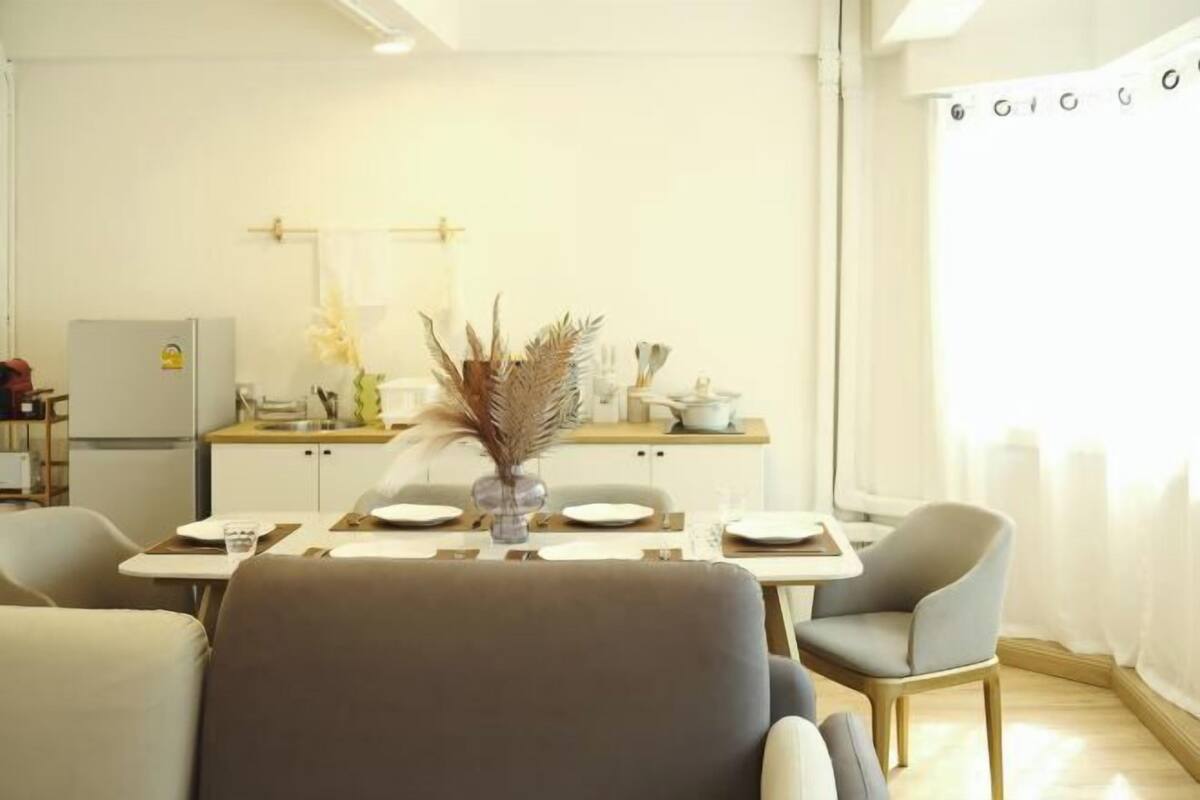 Ein Essbereich mit einem Tisch für vier Personen, eine Küche im Hintergrund und ein Fenster mit Vorhängen.