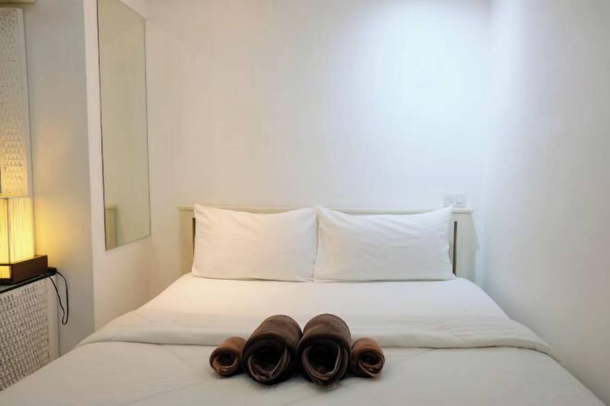 A hotel room with a bed, two pillows, a lamp, and rolled towels on the bed.