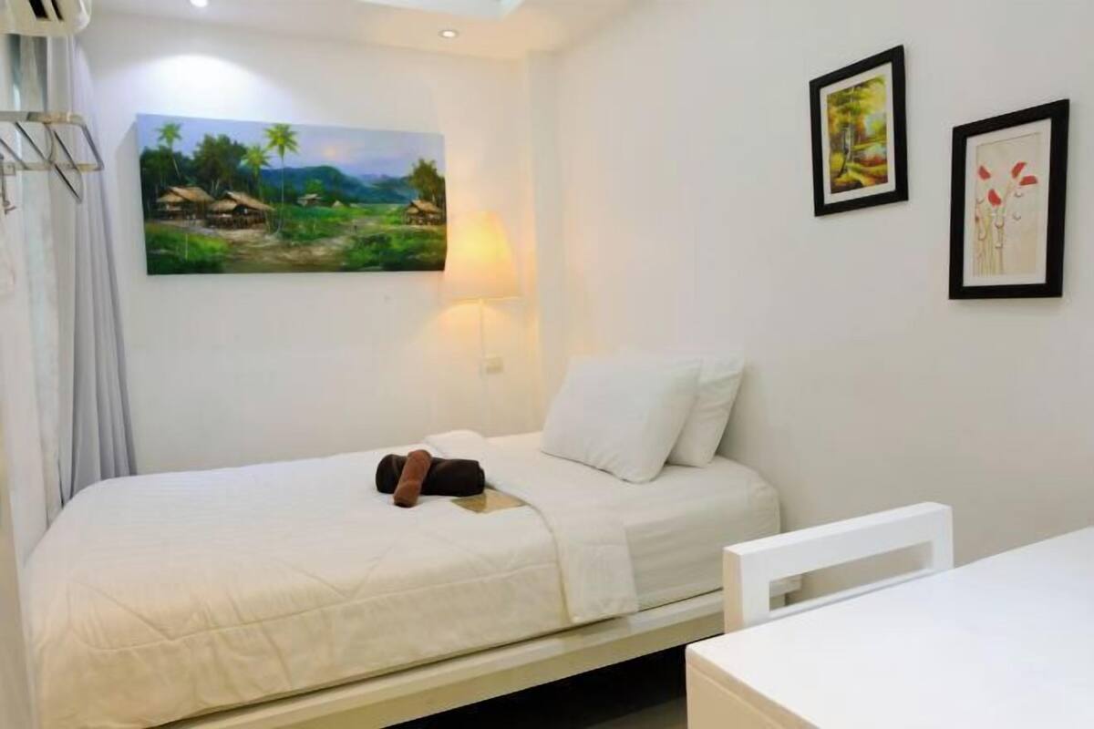 A single bed with a white mattress and pillows, a bedside table, a wall-mounted lamp, and two framed artworks on the wall.